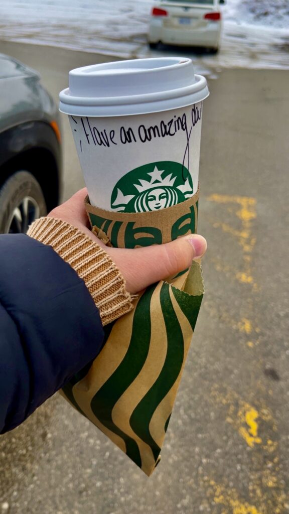 A hand holding a Starbucks takeaway coffee cup with a cardboard sleeve, featuring a handwritten message that reads “Have an amazing day,” outdoors in a parking lot with patches of snow and parked cars in the background.